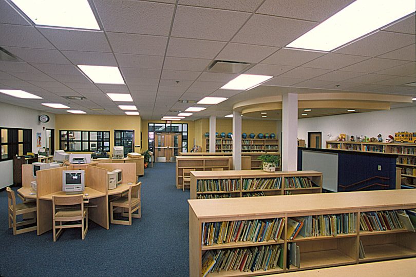 Craig Houghton Library with dark yellow accent wall and light wooden shelving and circular computer desks