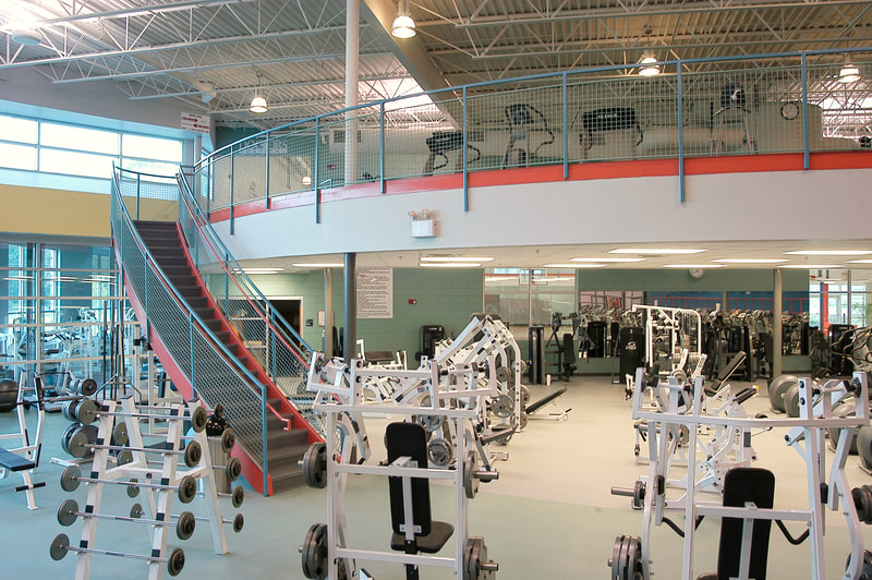 view of many weight machines and free weights on bottom floor with colorful blue and red staircase leading to an open air second floor with cardio machines and large windows at Augusta Family Y 